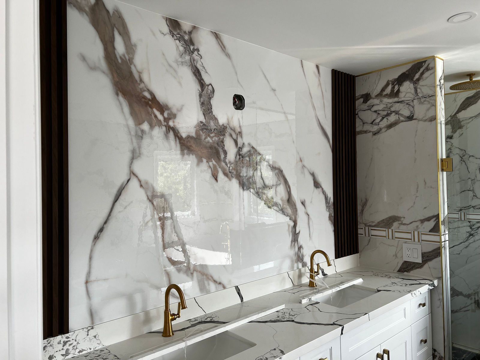 Modern bathroom vanity with marble walls, dual sinks, gold faucets, and a large mirror