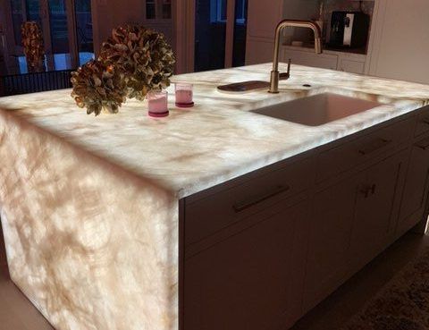 Sunlit kitchen island with a glowing stone countertop, sink, faucet, and a vase of dried flowers