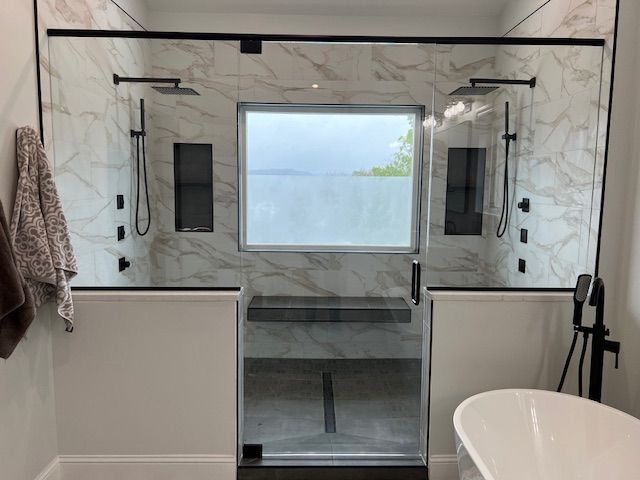 Modern bathroom with marble walls, large window, glass shower door, and freestanding tub