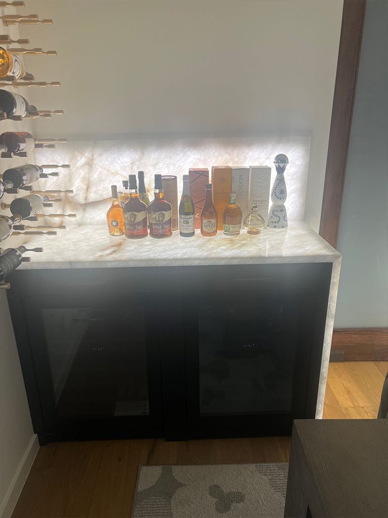 Black cabinet with marble top displaying several liquor bottles against a bright wall.