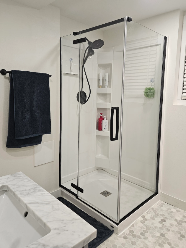 Modern glass shower enclosure with black trim in a bright bathroom, beside a white sink and dark towel