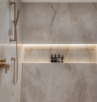 Minimalist beige shower with gold fixtures, wall niche, and lit shelf holding toiletries
