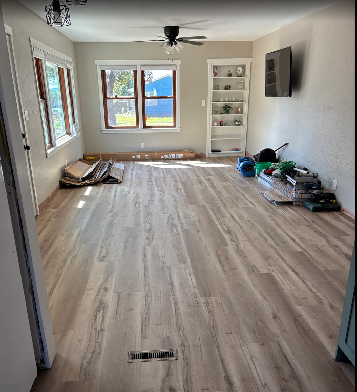 Bright living room with light wood floor, windows, white shelving, and cleaning supplies along the wall.