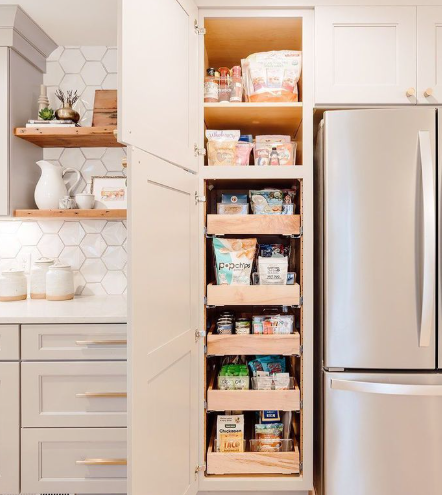 Open pantry beside a stainless steel fridge, with shelves stocked with jars, boxes, and baskets.