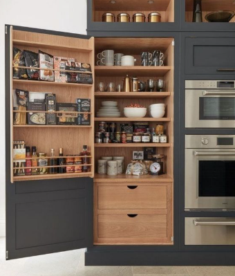 Open pantry cabinet with spices, jars, dishes, and drawers beside built-in stainless steel ovens.