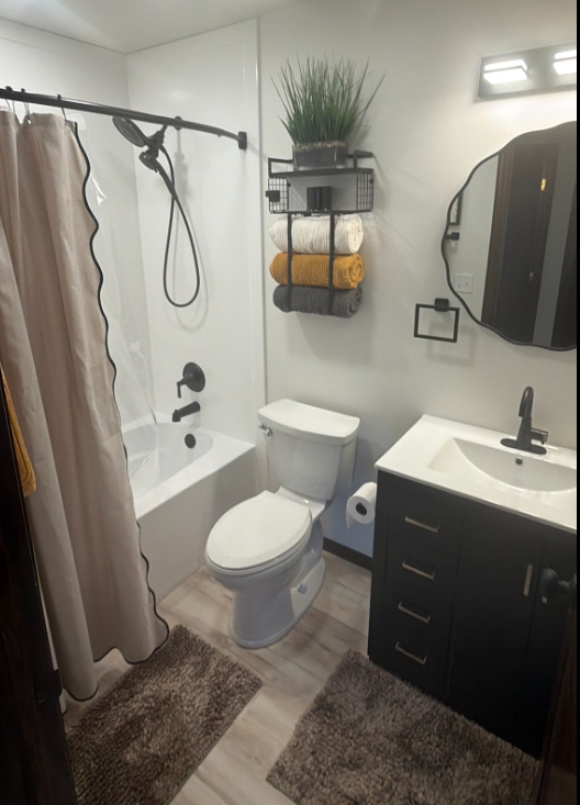 Small bathroom with bathtub, toilet, vanity, shower curtain, wall shelves, and rugs.
