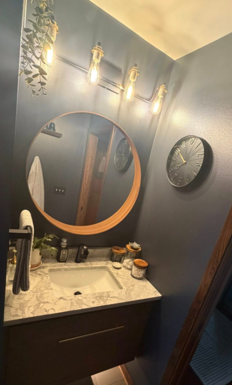 Small bathroom vanity with round mirror, wall lights, dark cabinet, and clock on a blue-gray wall.