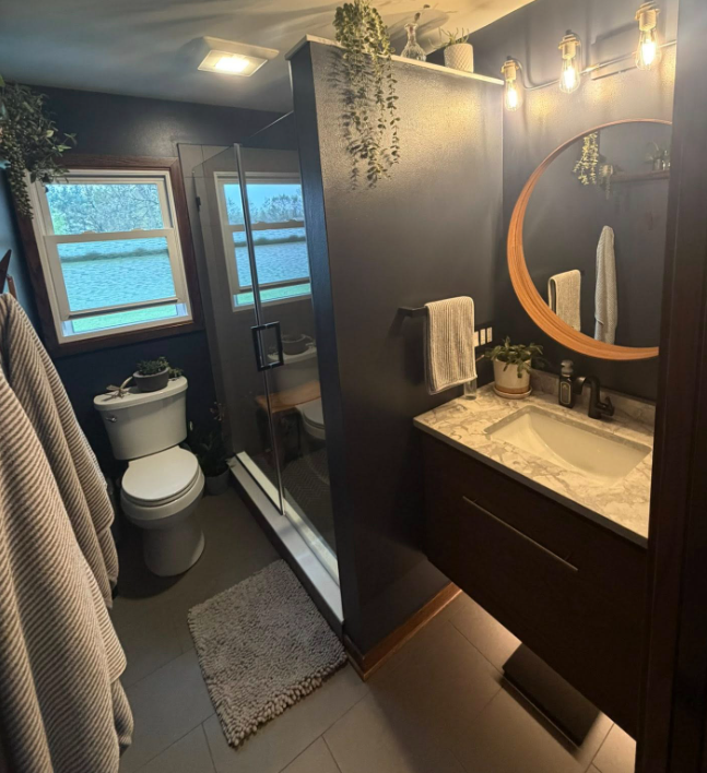 Compact modern bathroom with dark walls, round mirror vanity, toilet, shower, and natural light from windows