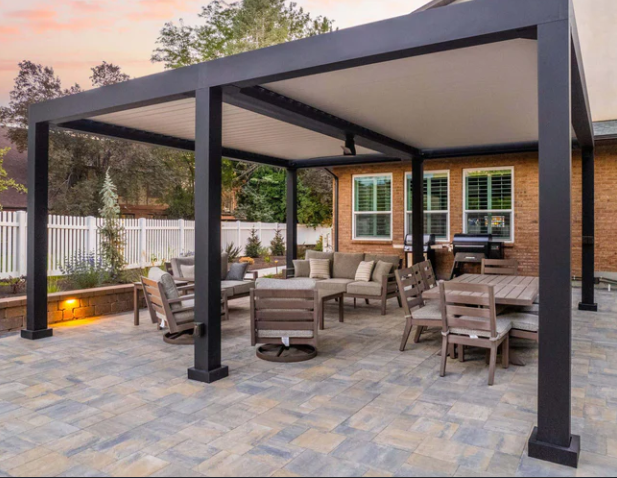 Covered patio with outdoor seating, a modern pergola, and stone pavers beside a house at sunset