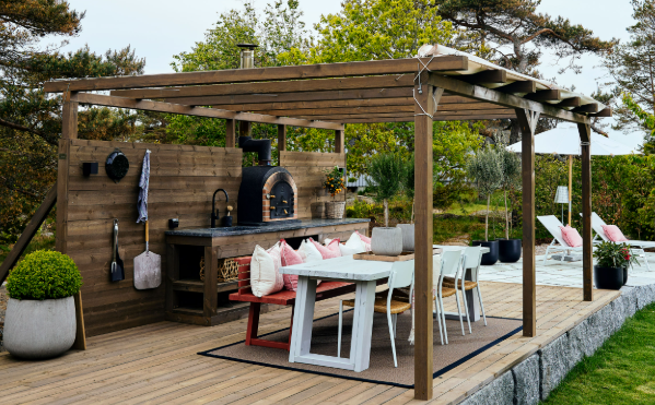 Outdoor wooden patio with a pergola, dining table, brick grill, and lounge chairs overlooking a garden