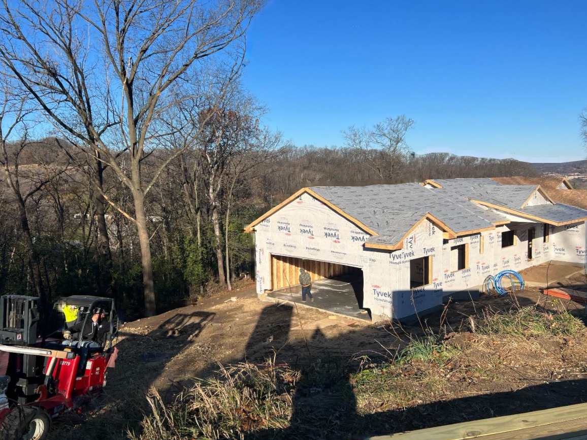 House under construction on a wooded hillside, with a red tractor in the foreground and clear blue sky.