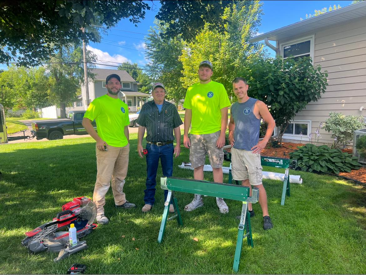 Four people stand in a sunny yard beside lawn tools and a green workbench.