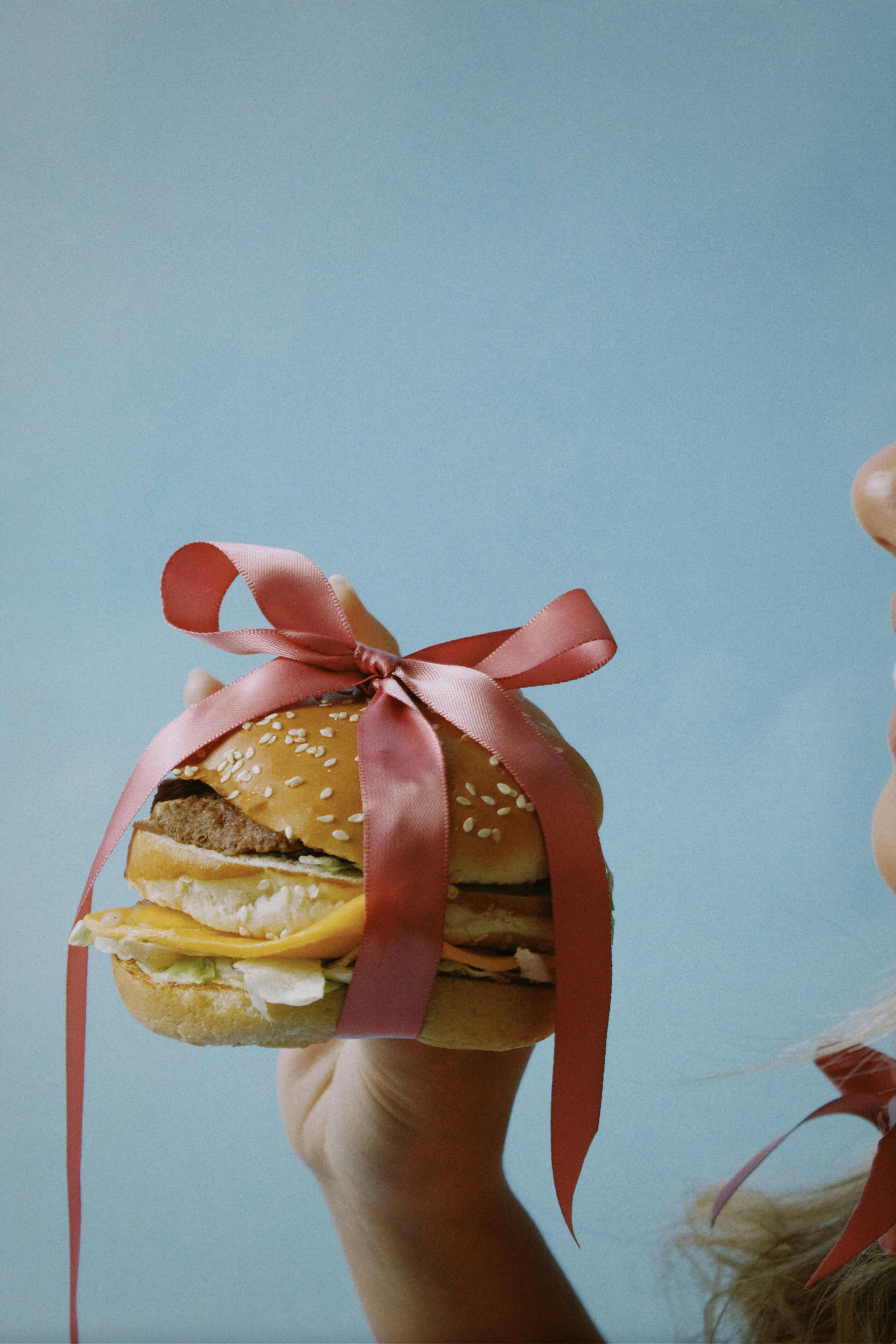 Burger wrapped with pink ribbon and held up against a blue background.