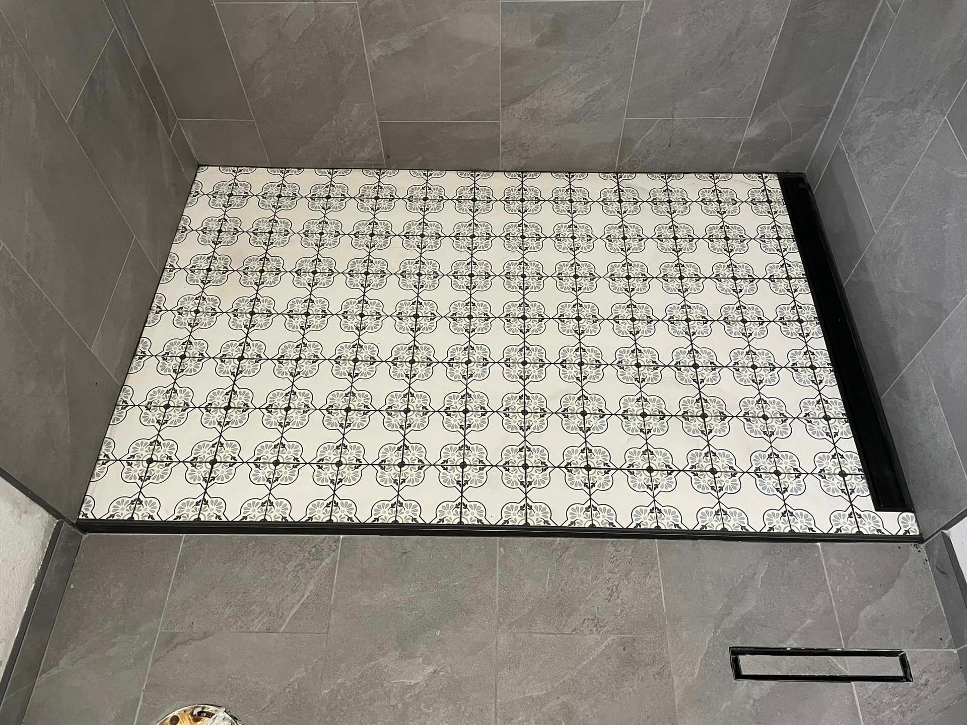 Gray tiled alcove with a white patterned mat on the floor and a small black handle on the lower right