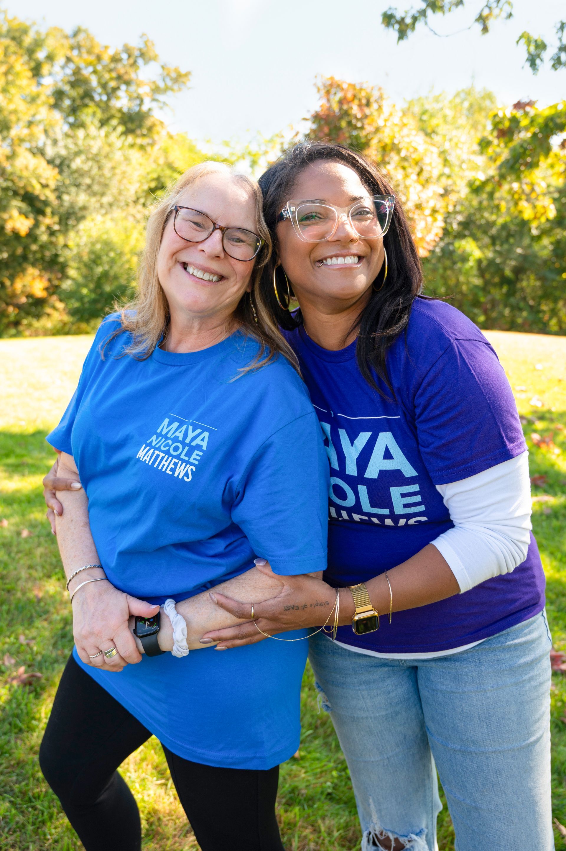Two smiling women embrace outside. Both wear blue shirts that say