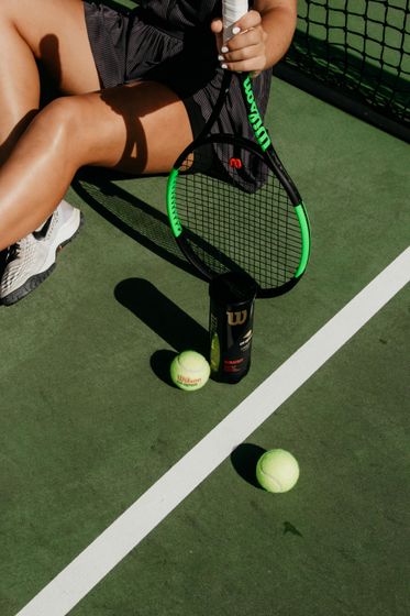 Woman sitting on a tennis court with a racket, tennis balls, and a can of balls.