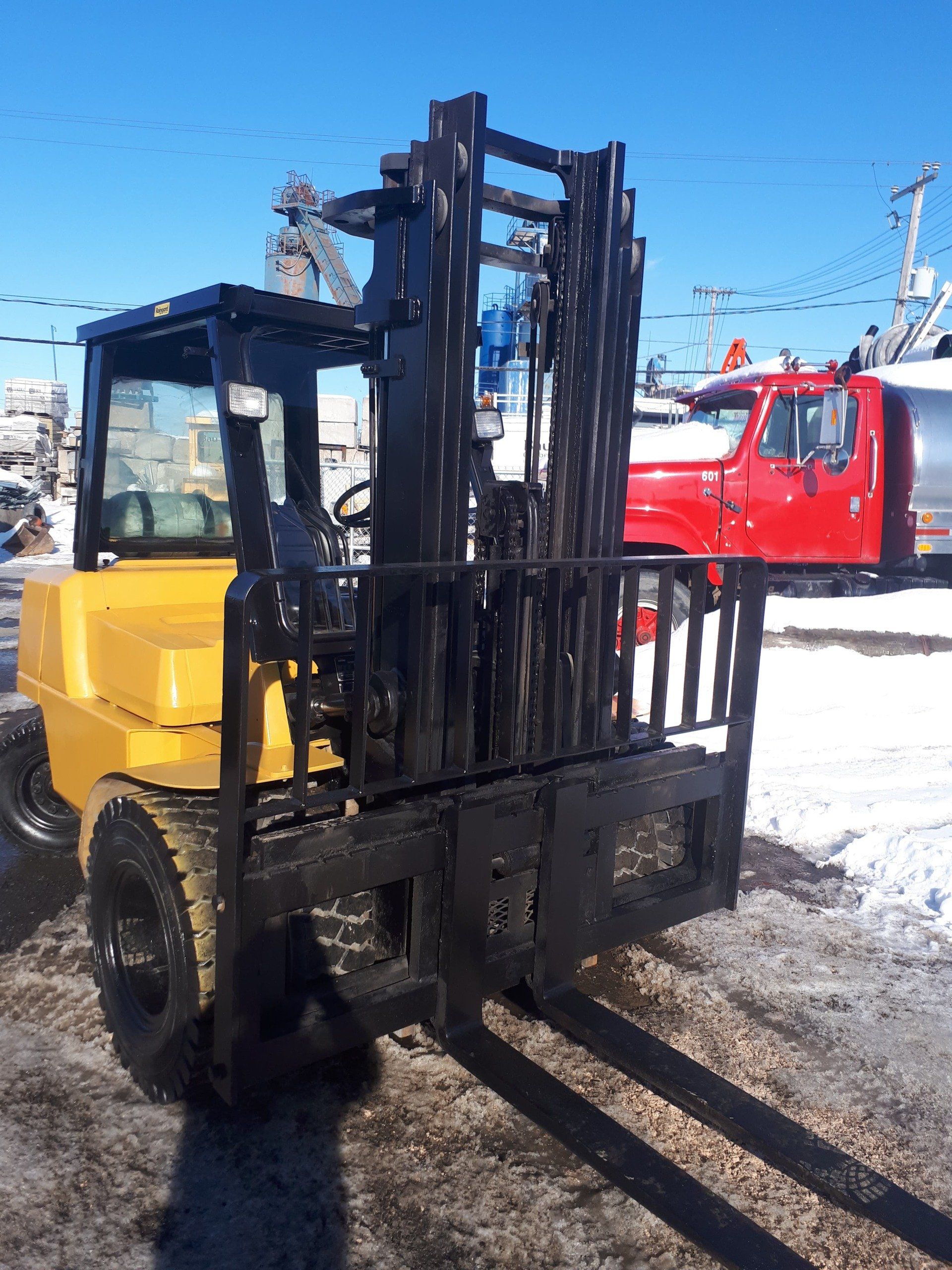 A yellow forklift is parked in the snow next to a red truck