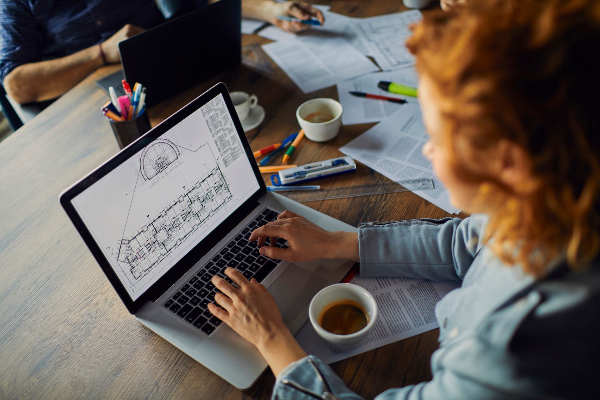 Une femme rousse travaille sur son ordinateur portable à une table, les yeux rivés sur un dessin. Une tasse de café et des papiers sont posés sur la table.