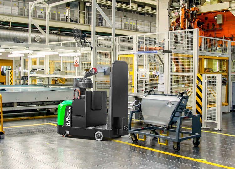 Véhicule à guidage automatique (AGV) remorquant un chariot métallique avec une porte de voiture dans un environnement d'usine.