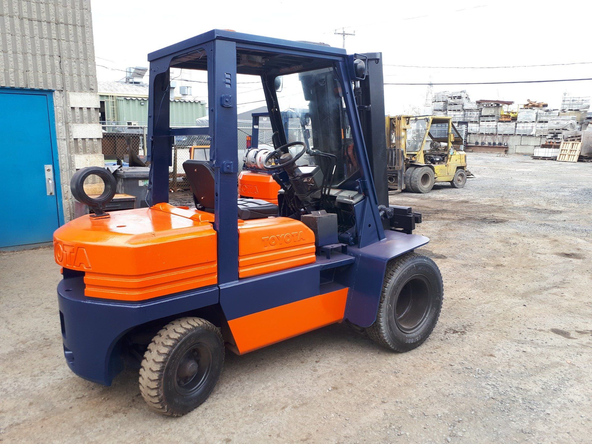 Un chariot élévateur bleu et orange sur un parking goudronné, avec un autre chariot élévateur en arrière-plan.