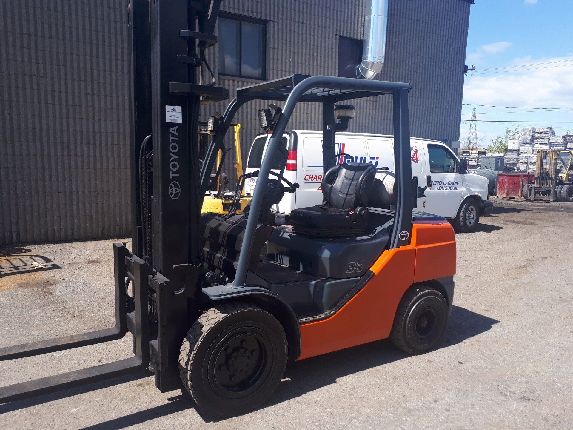Chariot élévateur Toyota orange sur une surface pavée à côté d'un bâtiment et d'une camionnette blanche.