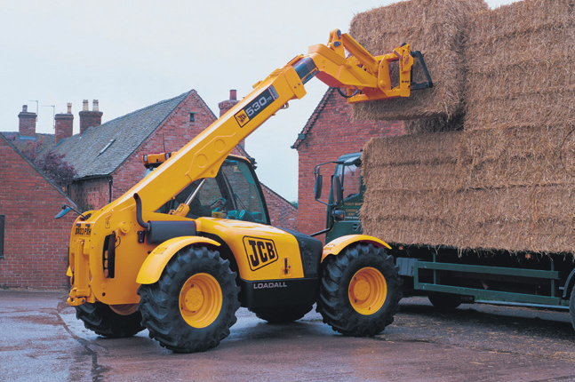 Un chariot télescopique JCB jaune charge des balles de foin sur un camion.