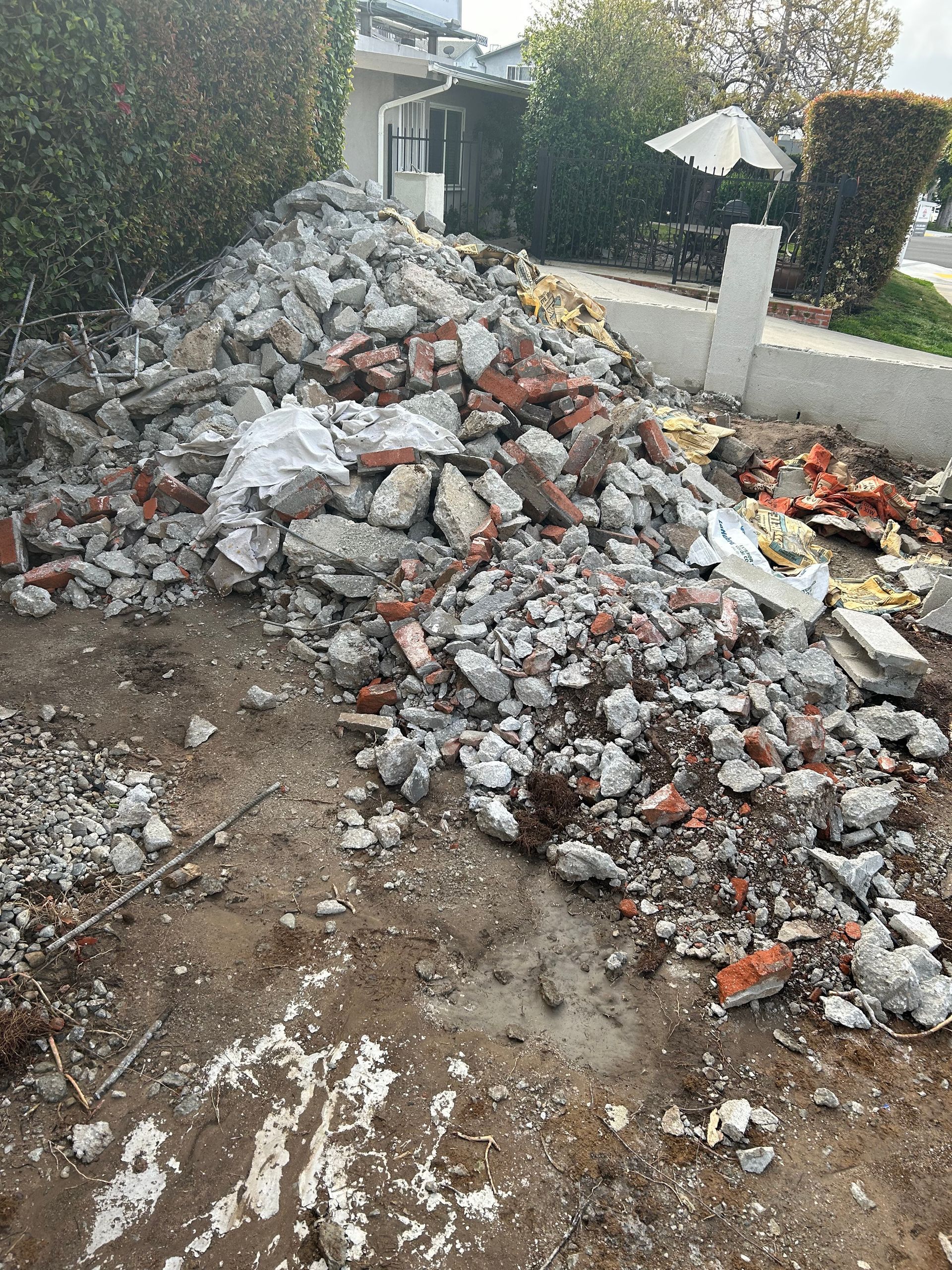 A large pile of broken concrete, bricks, and construction debris sits on dirt in a residential yard near a hedge.