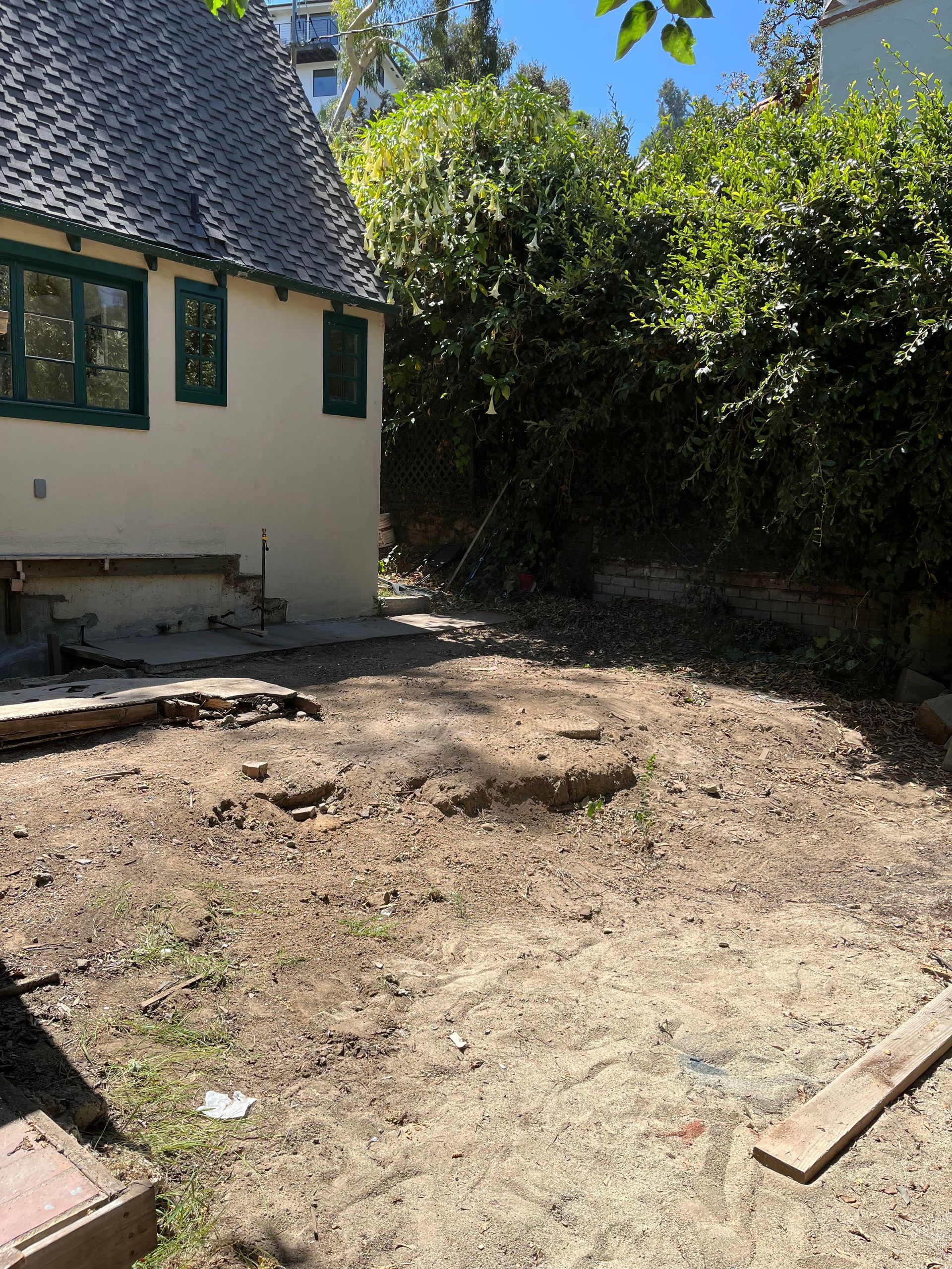 A side view of a cream-colored house with green trim next to a bare, dirt-filled backyard with trees in the background.