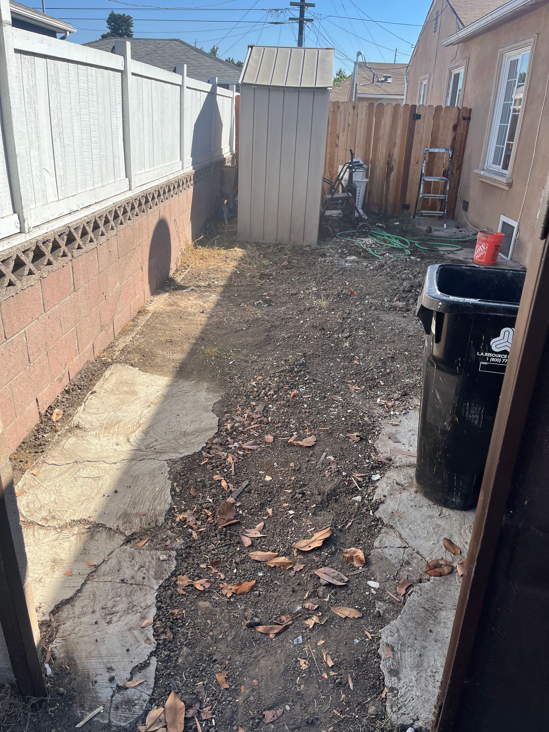 A narrow, debris-covered concrete path runs alongside a tan house and a brick wall, leading toward a metal storage shed.