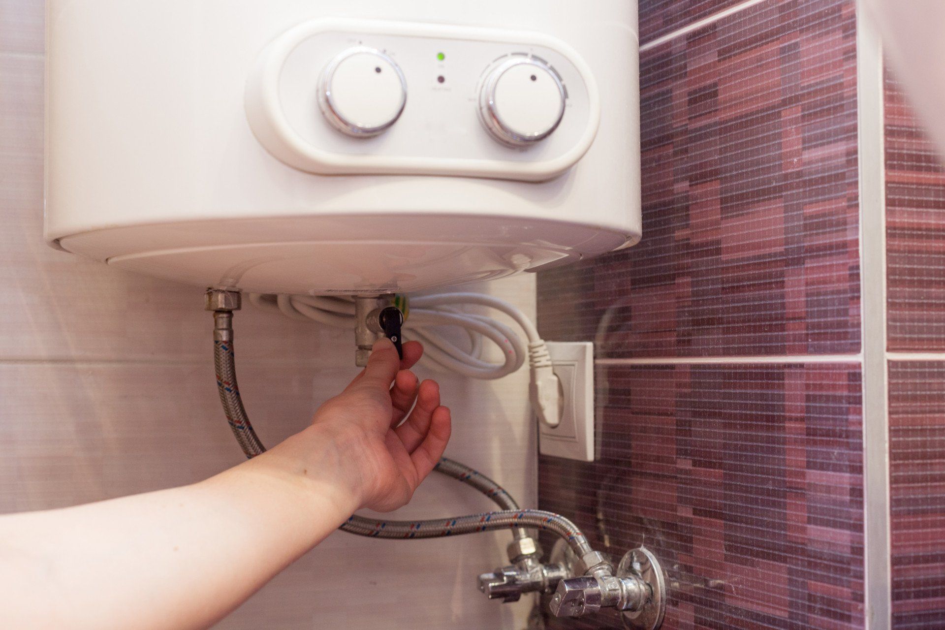 Hand turning a water heater valve under a white wall-mounted unit in a tiled bathroom