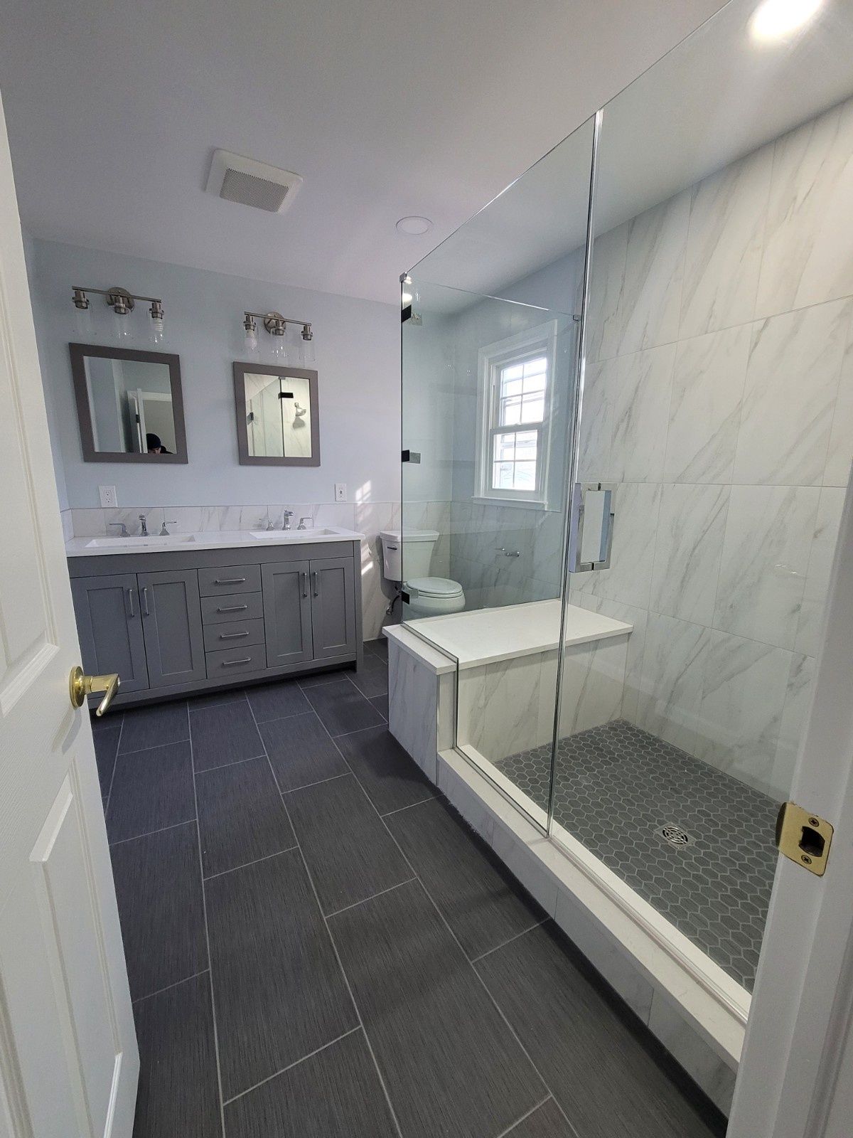 A modern bathroom featuring a gray double vanity, large floor tiles, and a walk-in glass shower with a tiled bench seat.