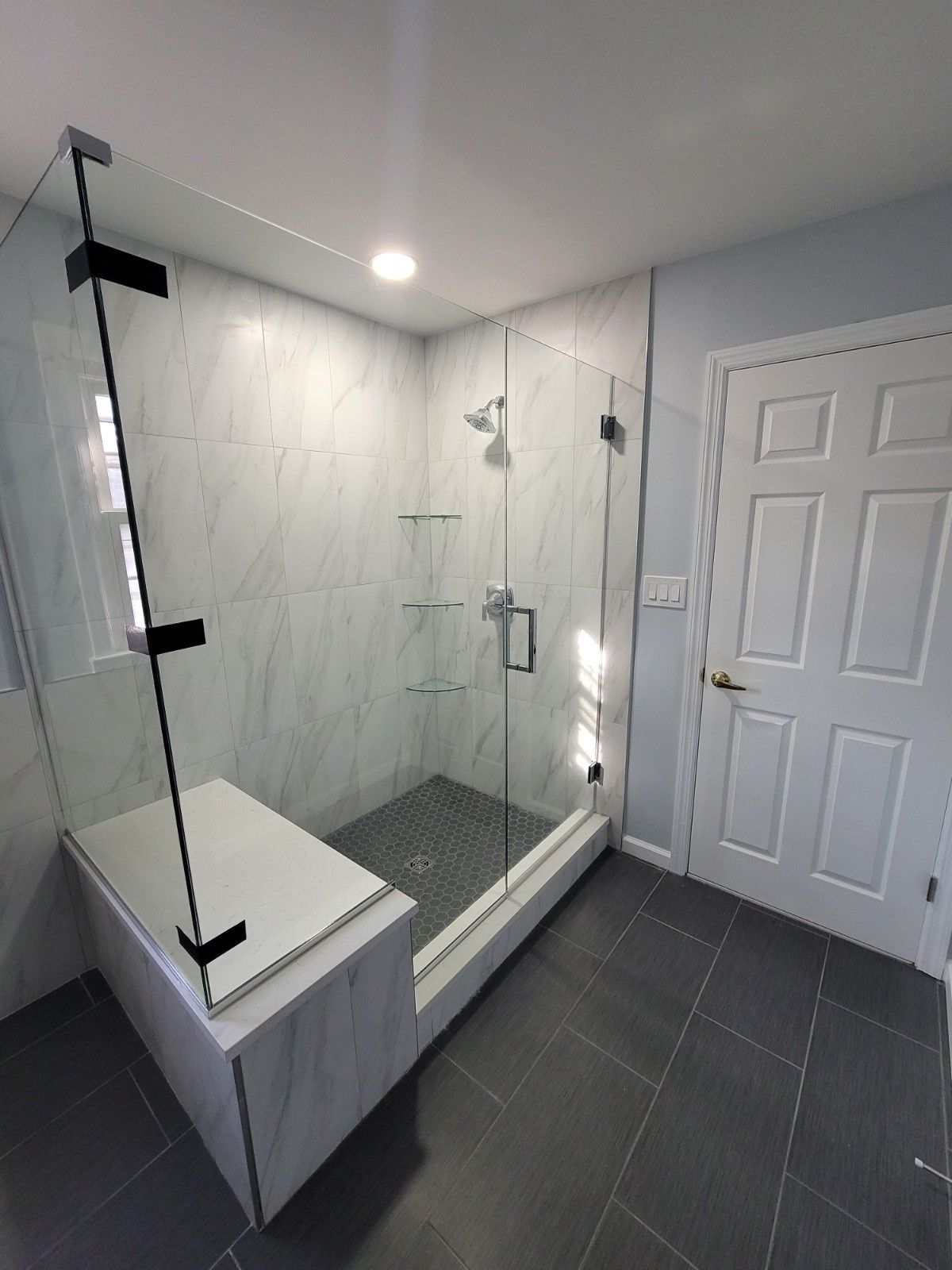 A modern walk-in shower with white marble-patterned tile, a dark gray tiled floor, glass panels, and a built-in bench.