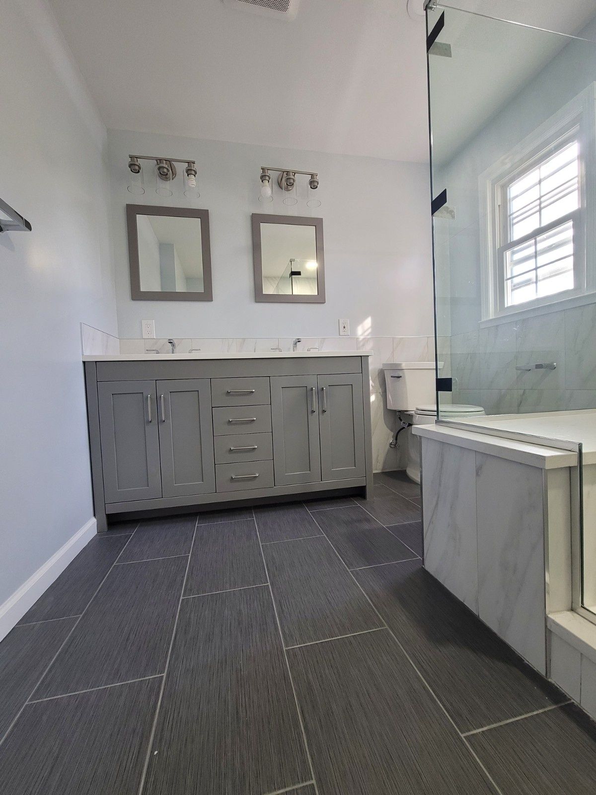 A modern bathroom with a double-sink grey vanity, two framed mirrors, a toilet, and a glass-enclosed shower on grey tiled floor.