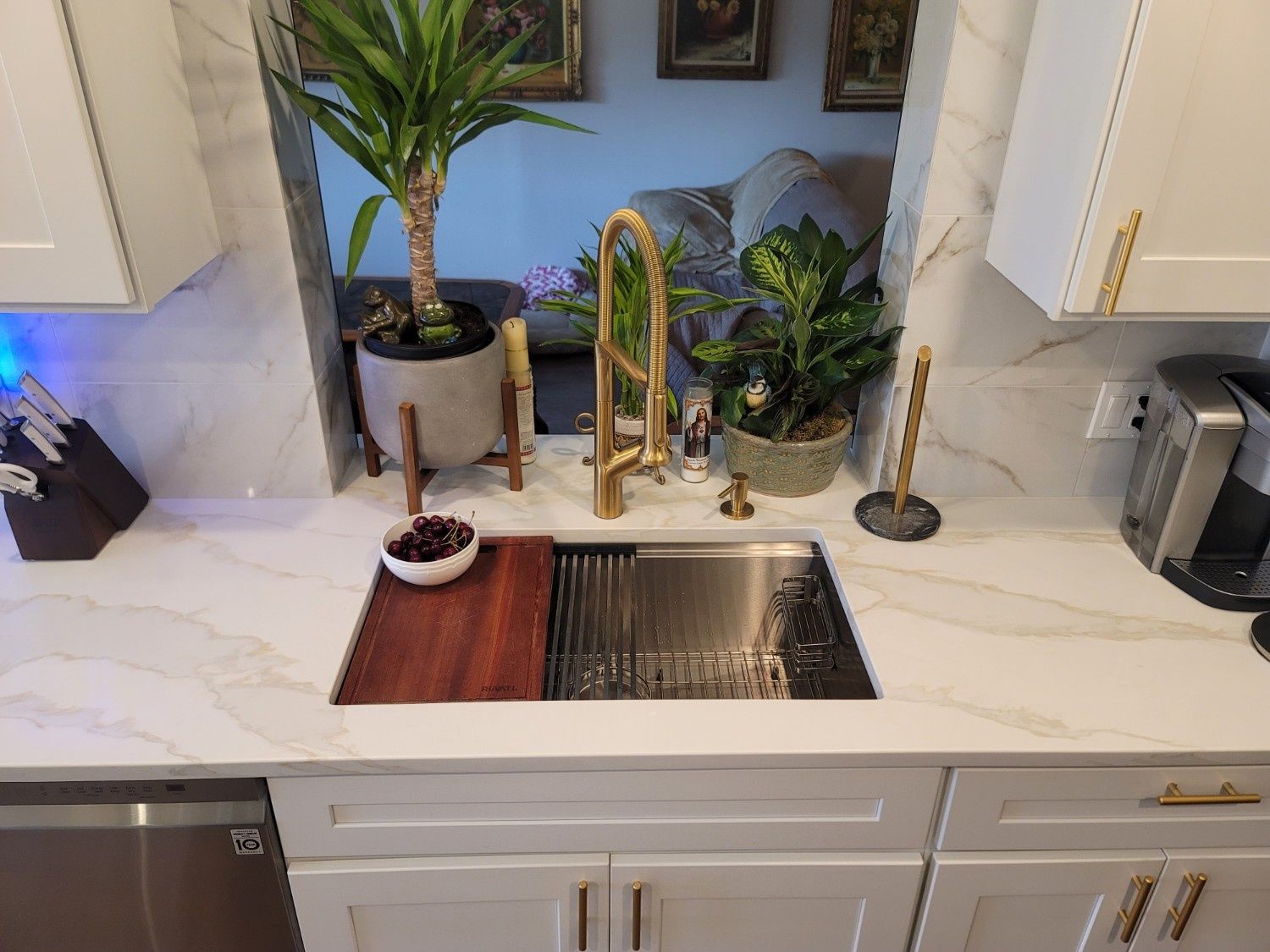 A white kitchen sink with a gold faucet, a wooden cutting board, a drying rack, and several potted plants on the counter.