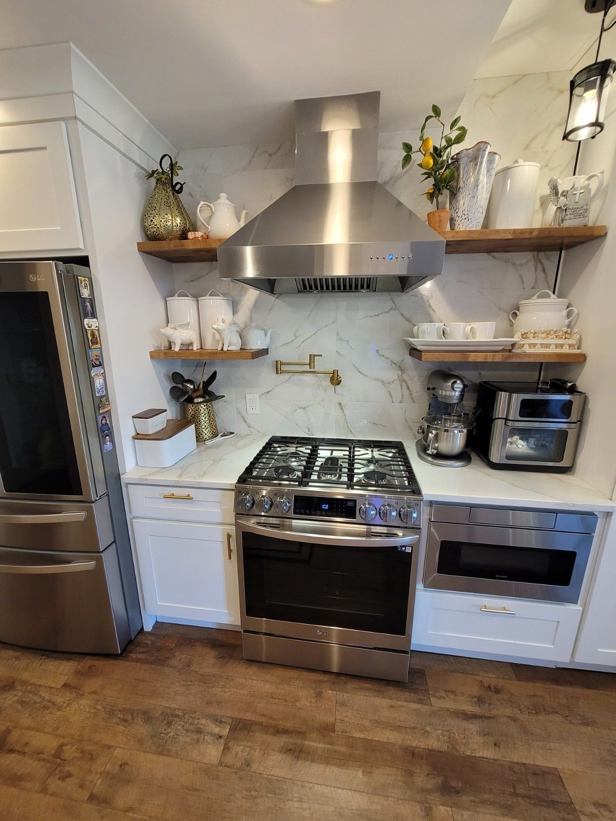 A bright kitchen featuring a stainless-steel stove, range hood, and microwave, with white cabinetry and wood shelving.