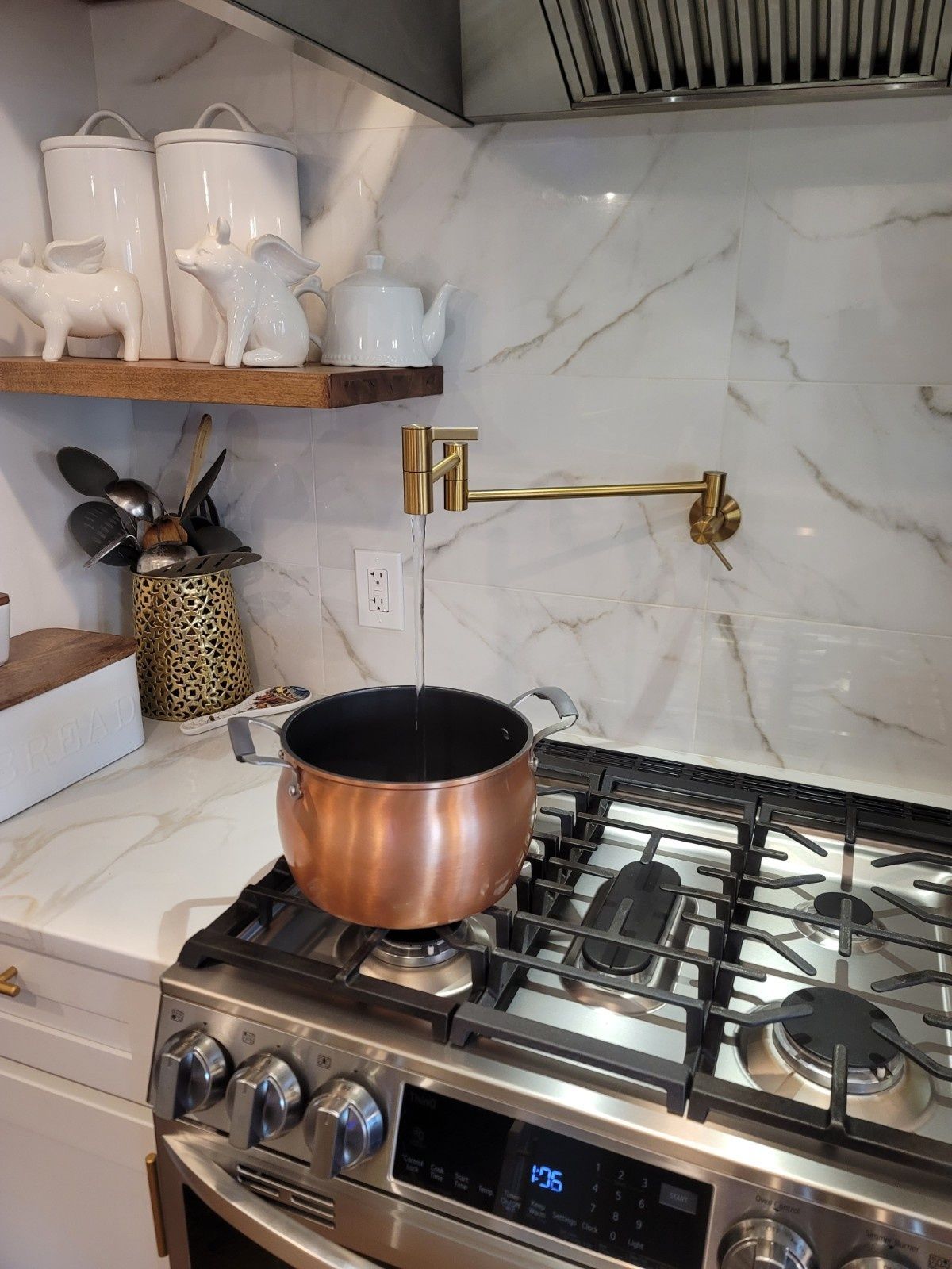 A copper pot sits on a gas stove under a gold wall-mounted pot filler faucet in a kitchen with a white marble backsplash.