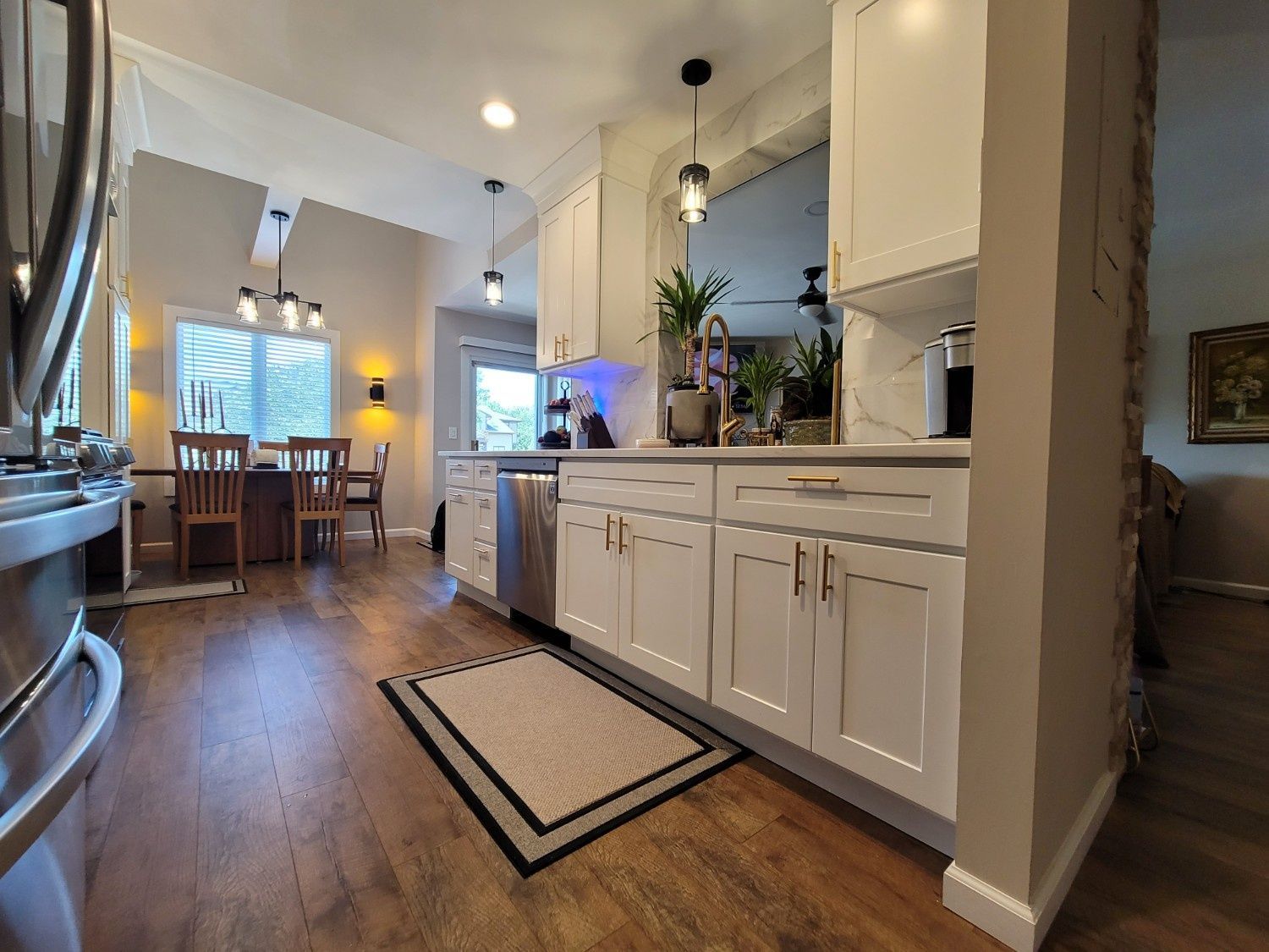 A modern white kitchen with stainless steel appliances, wood floors, and a dining area in the background.