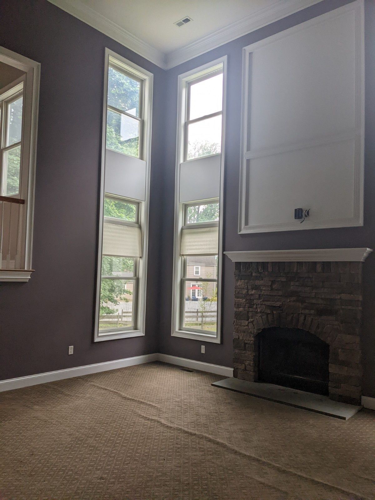 A living room with dark purple walls, beige carpet, tall windows, and a stone fireplace with a white-framed wall feature.