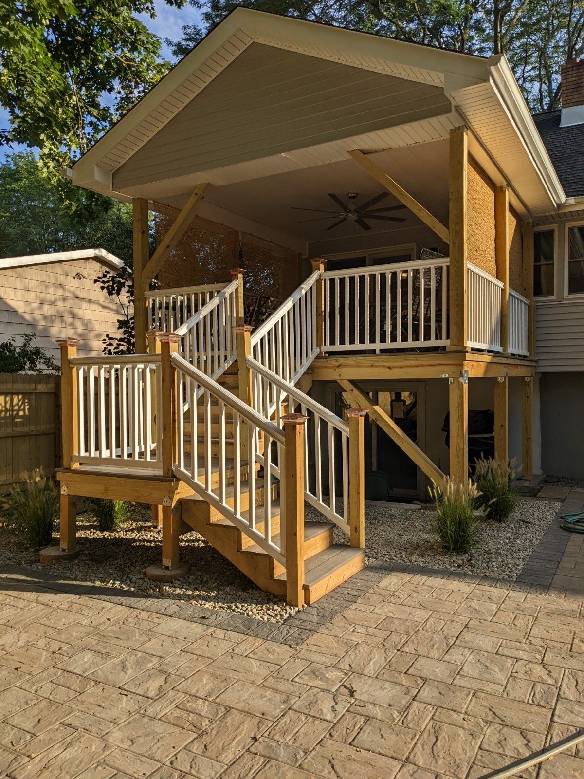 A newly constructed elevated wooden deck with white railings and stairs, positioned above a stone patio in a backyard.