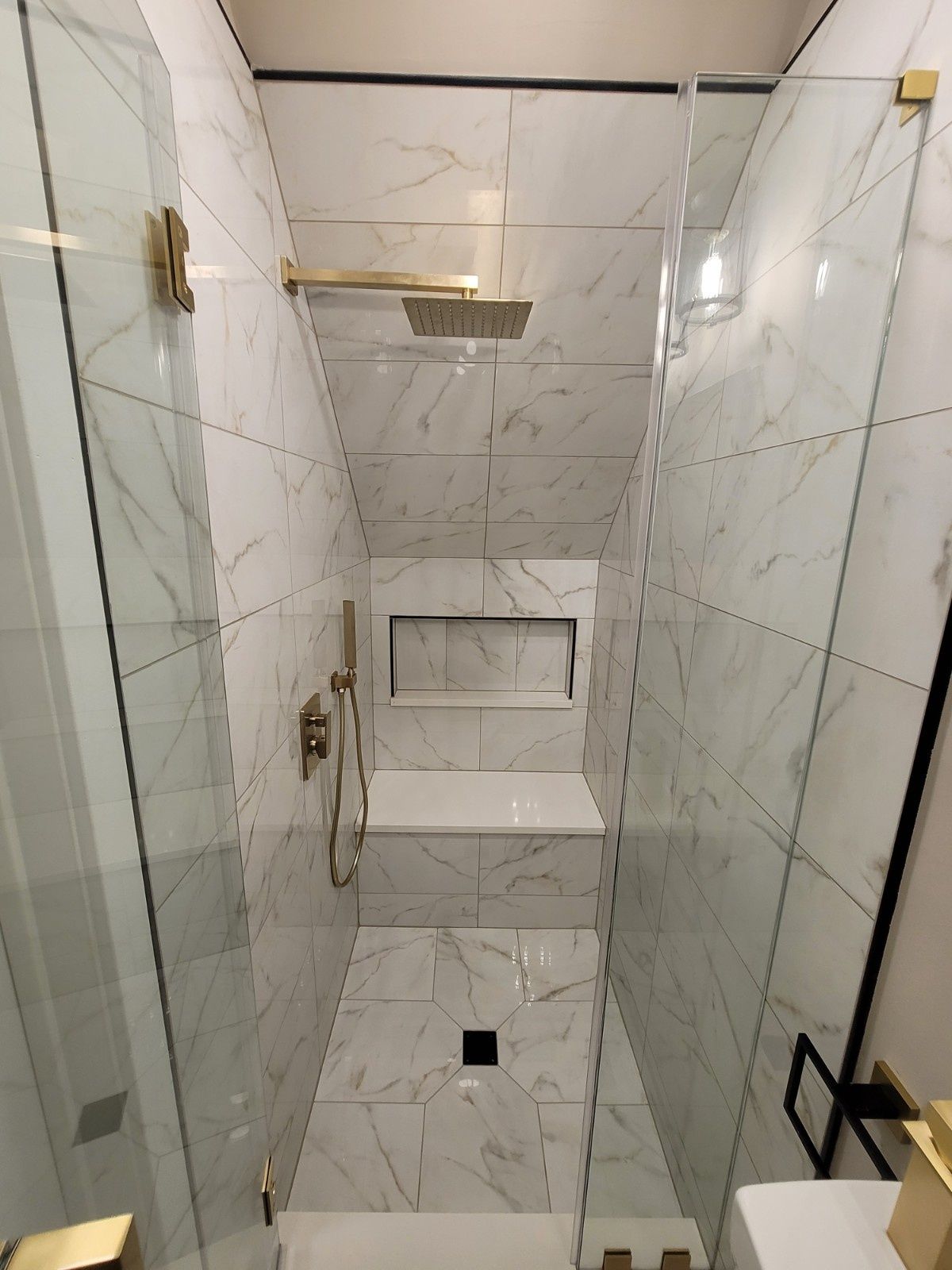 A marble-tiled walk-in shower with a built-in bench, gold fixtures, and a glass partition in a contemporary bathroom.