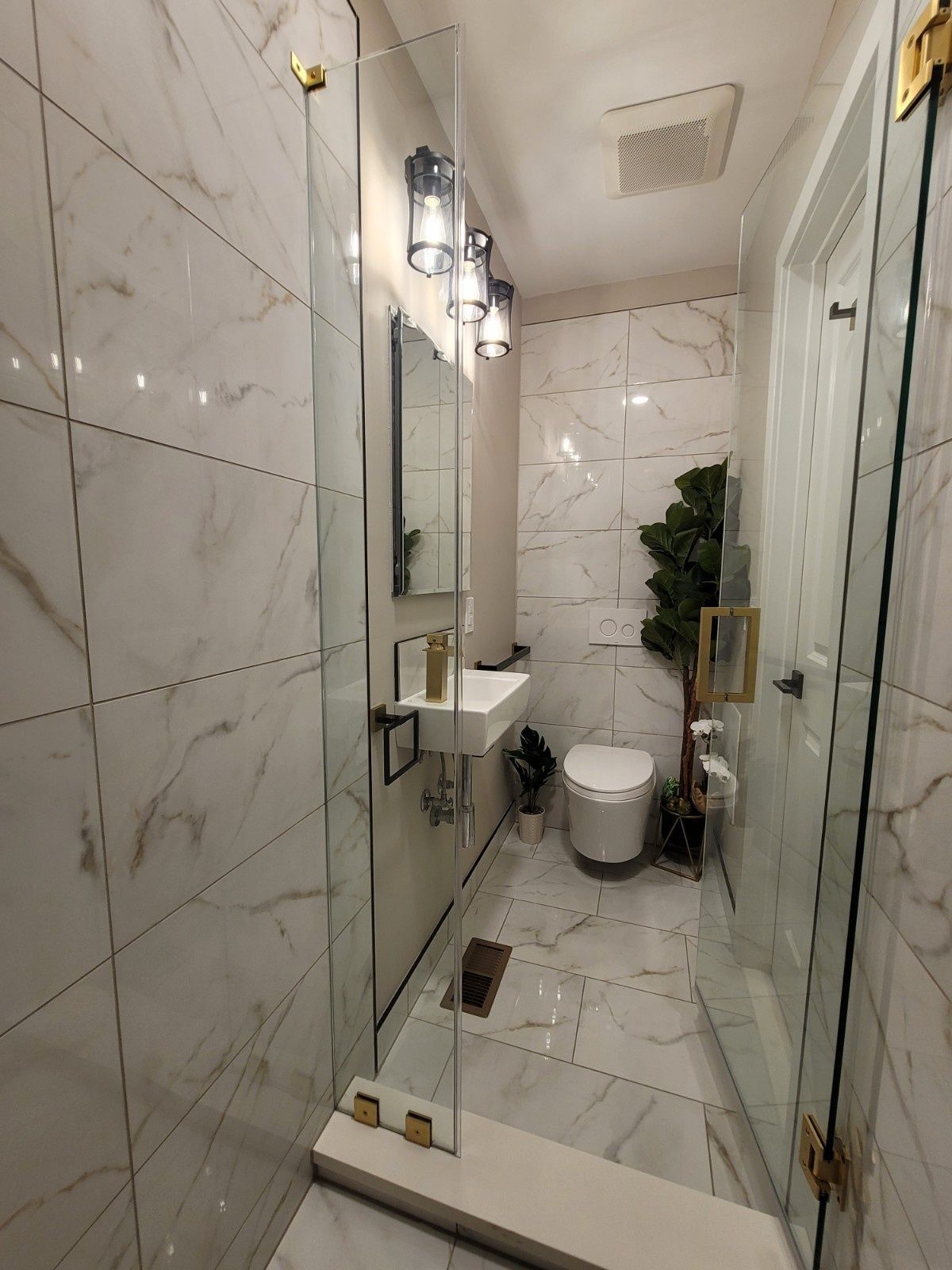 A narrow modern bathroom featuring white marble-patterned tiles, a glass shower door, a small vanity, and a toilet.