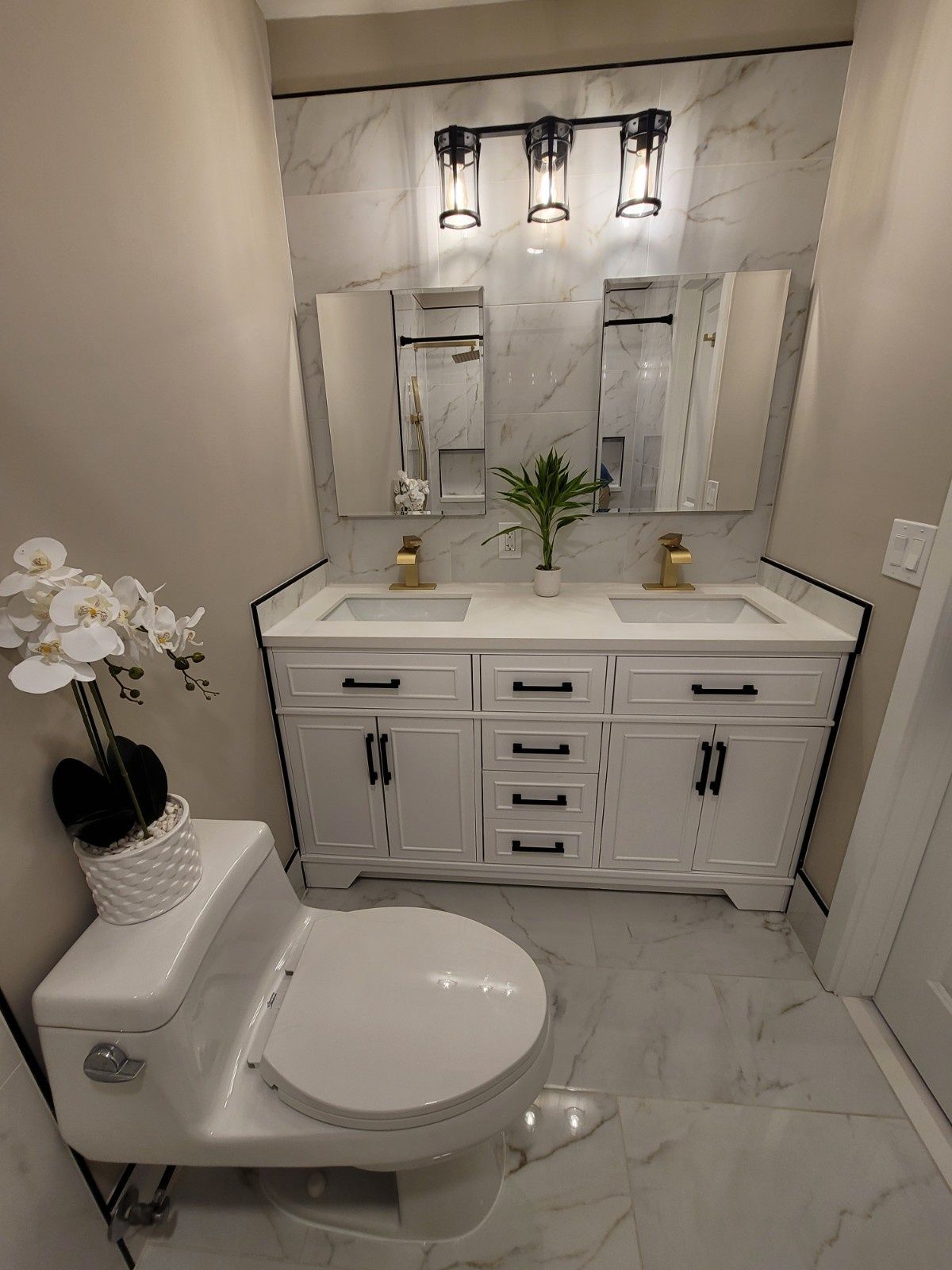 A white double-vanity bathroom with marble-style tile, gold faucets, black hardware, and a white toilet.