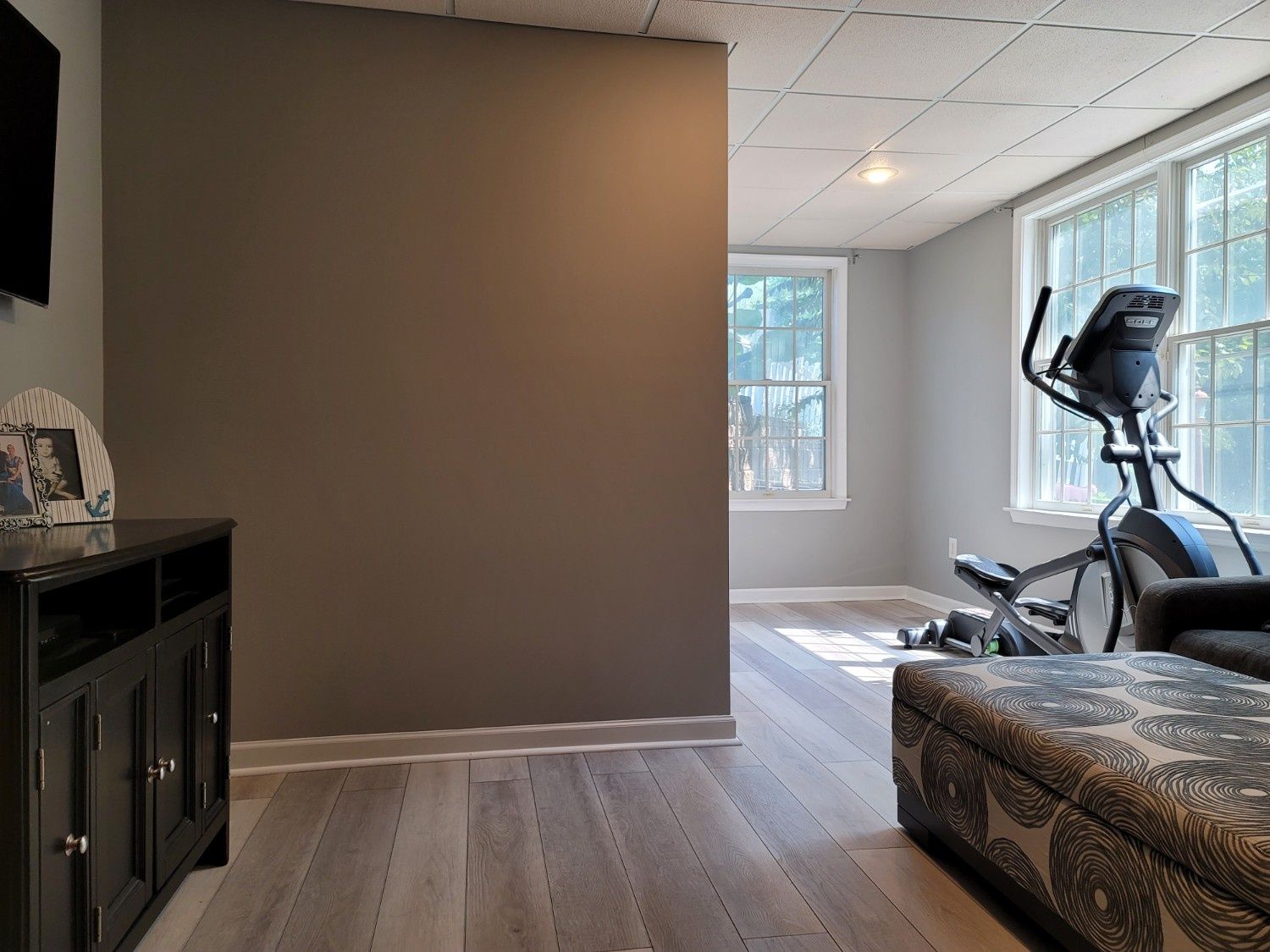 A brown accent wall in a room featuring light wood floors, a dark media console, and an elliptical machine by a window.