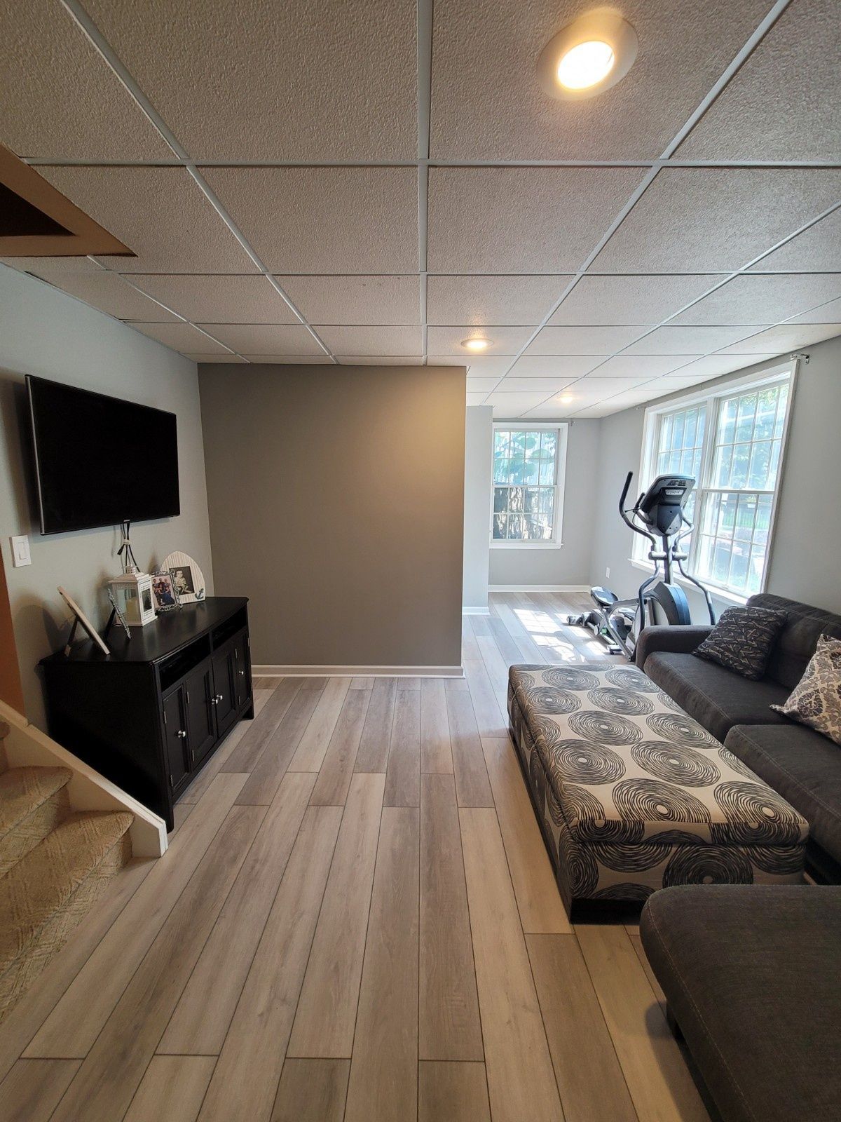 A basement living area with wood-look flooring, a TV on a black console, a gray sofa with a patterned ottoman, and gym gear.