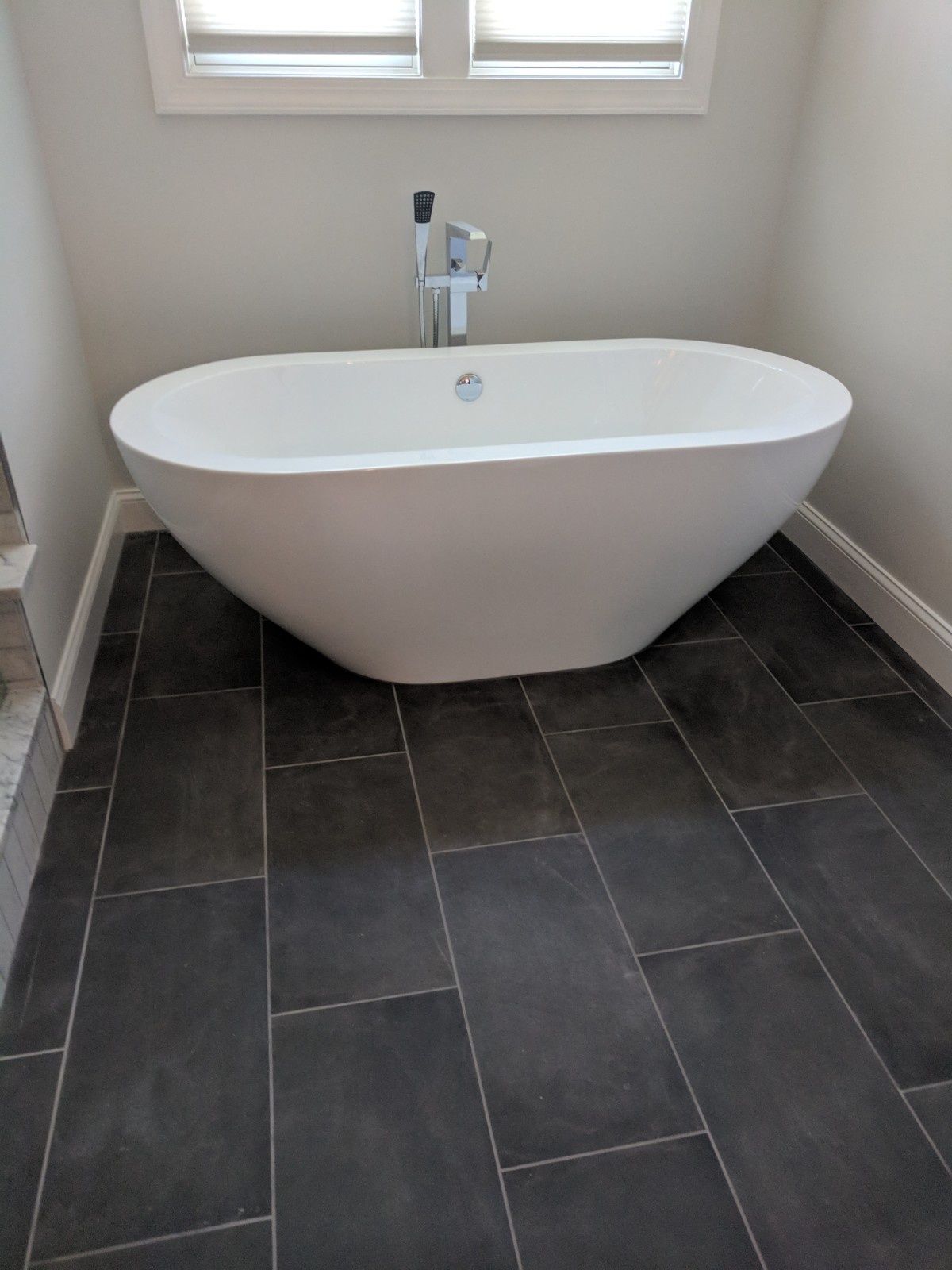 White freestanding bathtub with a silver faucet in front of a window, set on dark gray rectangular floor tiles.