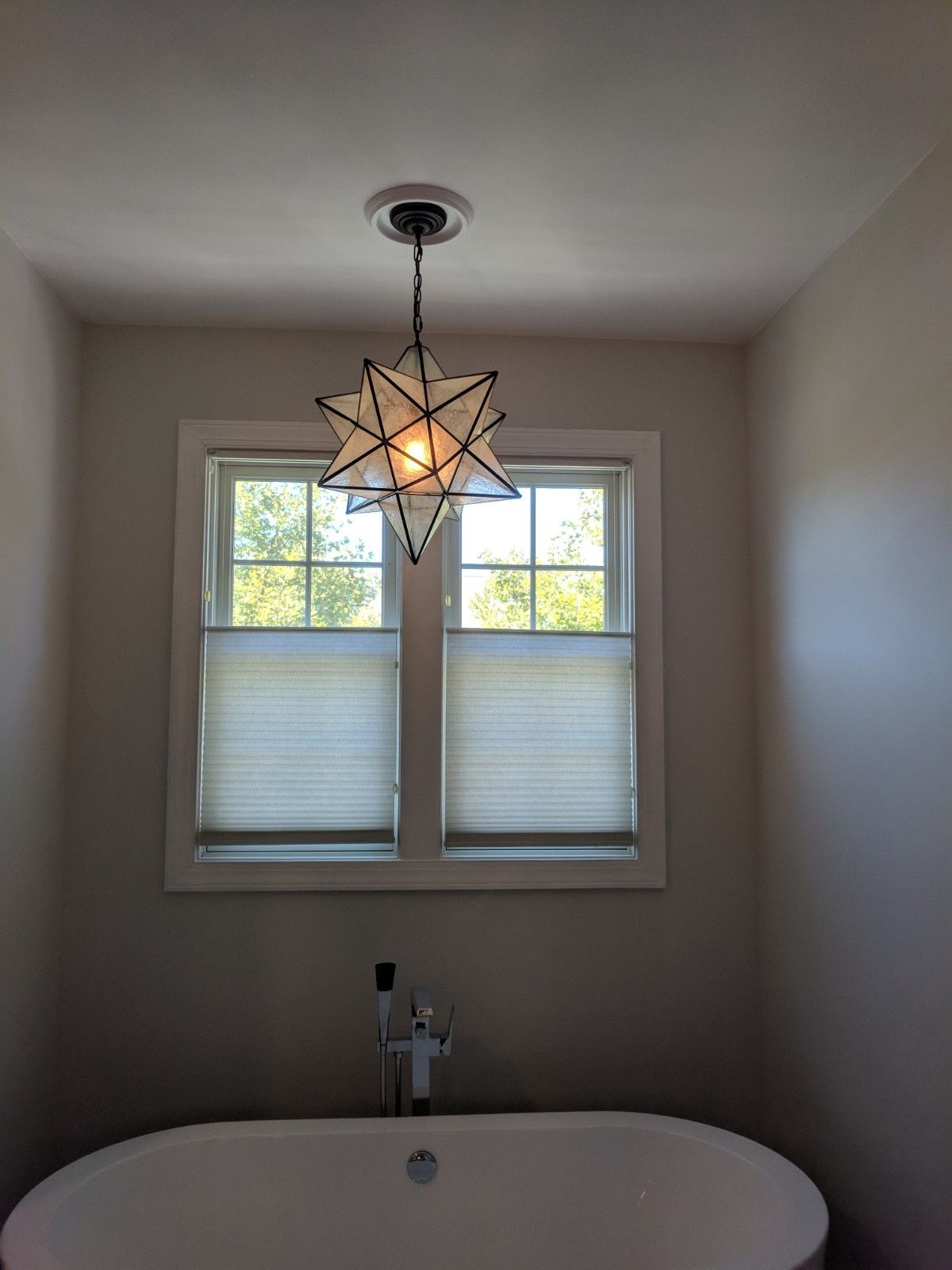 A star-shaped pendant light hangs above a freestanding white bathtub in front of a window with closed shades.