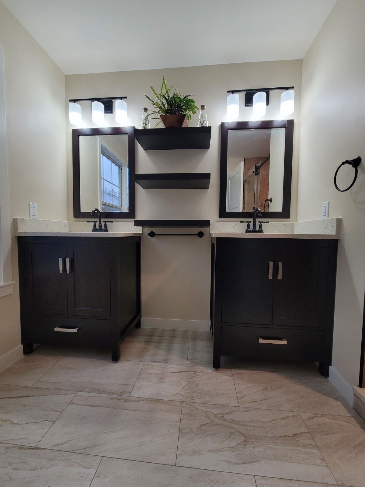 A double vanity bathroom with dark cabinets, two mirrors, wall lighting, and three floating shelves on a stone tile floor.