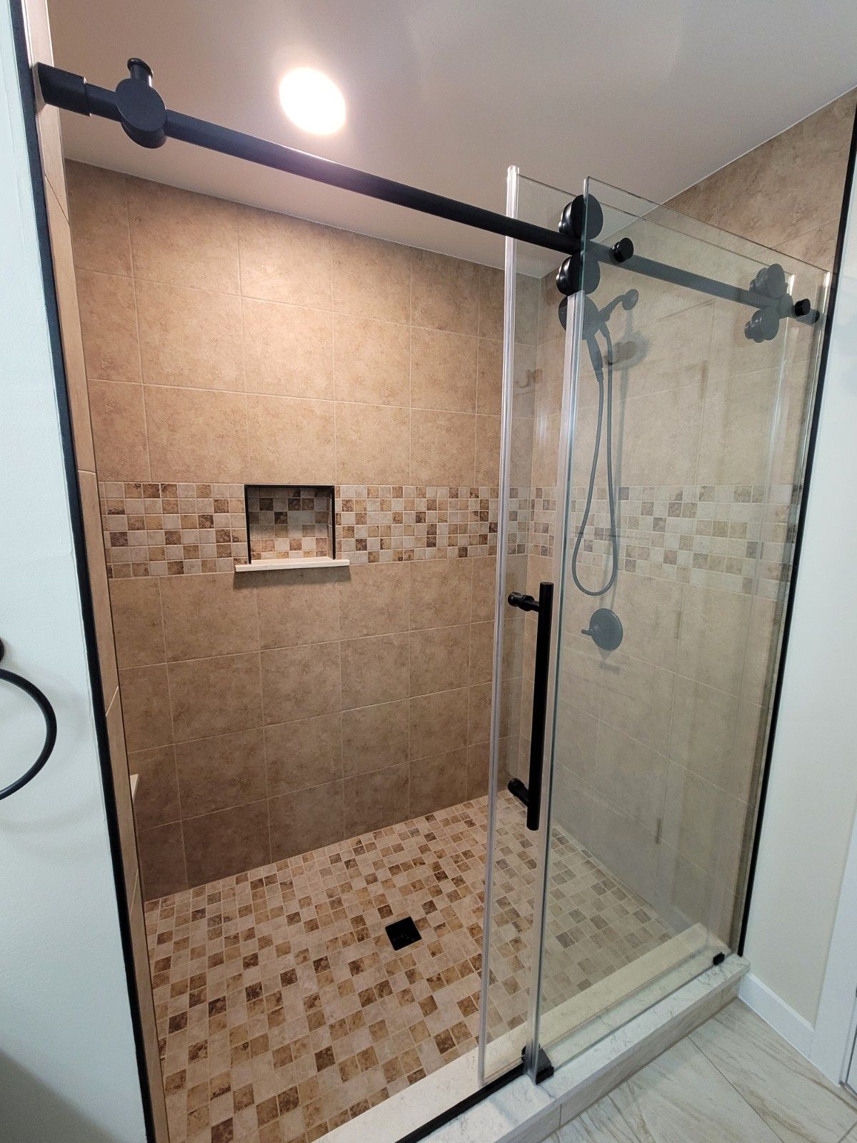 A walk-in shower with beige patterned tiles, a glass sliding door with black hardware, and a small built-in wall niche.
