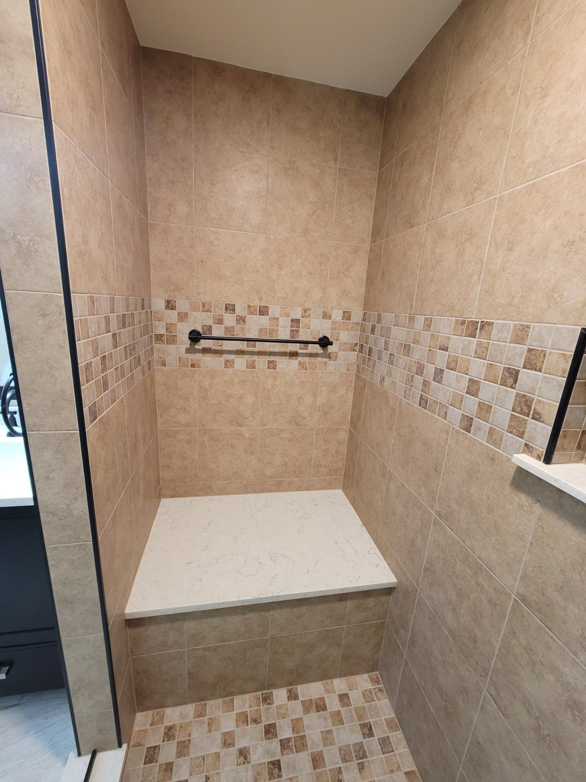 A shower stall featuring beige wall tiles, a mosaic tile accent strip, a white stone bench, and a dark metal towel rack.