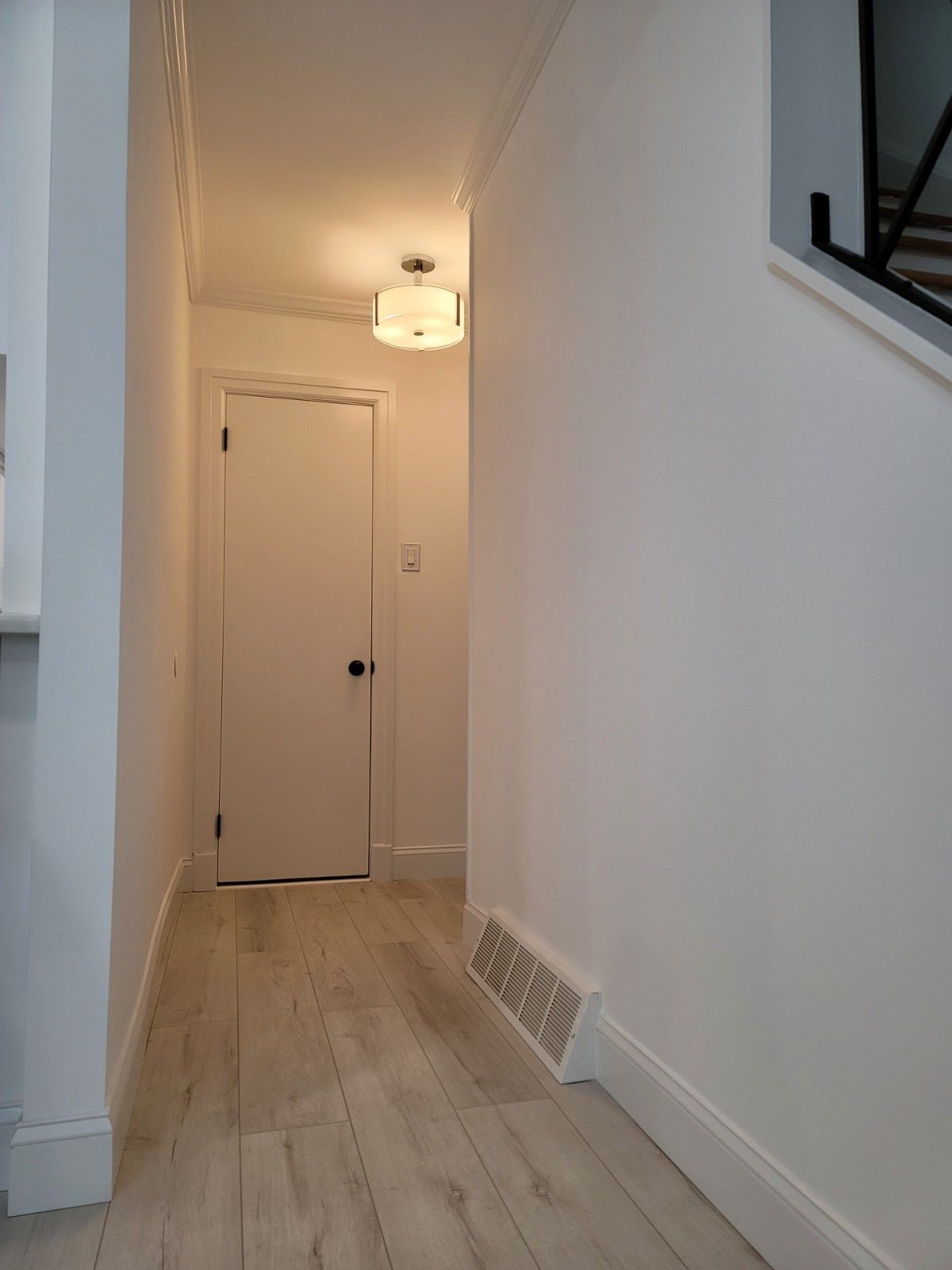 A brightly lit hallway with light wood floors, white walls, a white closed door at the end, and a modern ceiling fixture.